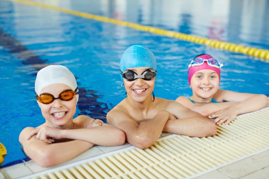 Three healthy smiling children looking to camera wearing swimming goggles and caps in water at tiled border of blue clearwater swimming pool